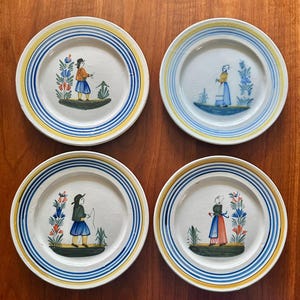 May include: A set of four white ceramic plates featuring hand-painted figures in traditional clothing and floral motifs. Each plate is decorated with concentric yellow and blue circular bands along the rim and a central illustration of a person.