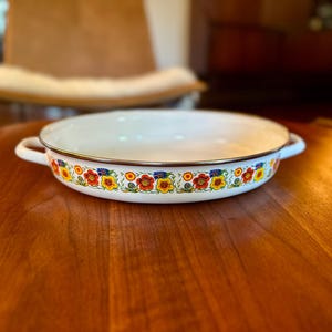May include: A shallow white enamel pan with two side handles and a metallic rim. The exterior features a colorful repeating pattern of red, yellow, and blue flowers with green leaves and stems encircling the entire circumference of the vessel.