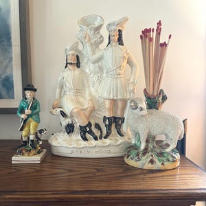 May include: A ceramic figurine of a person in a green coat and yellow breeches with a dog, a large cream-colored ceramic Robin Hood display piece with two figures and a dog, and a ceramic sheep figurine holding a bundle of long wooden matches.