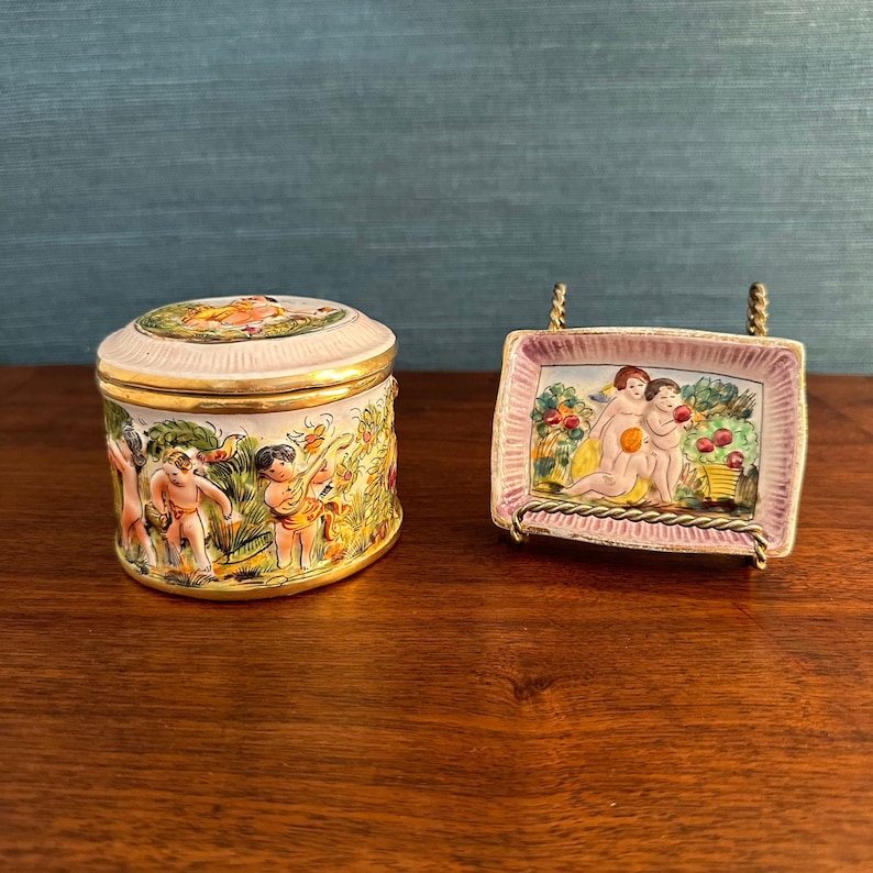 Puede incluir: Dos cajas de cer&aacute;mica para baratijas con escenas pintadas a mano de querubines en un jard&iacute;n. Las cajas est&aacute;n decoradas con detalles dorados y tienen un esquema de color rosa y blanco.
