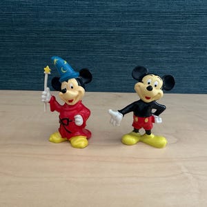 May include: Two Mickey Mouse figurines. One is dressed as a sorcerer in a red robe and blue hat, holding a wand. The other Mickey Mouse is in his classic outfit: black shirt, red shorts, and yellow shoes. Both figures are on a wooden surface.