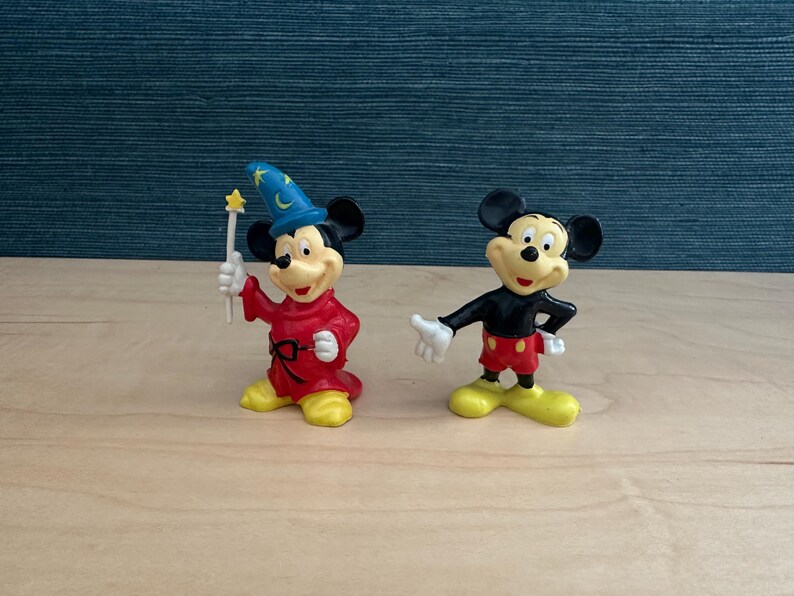 May include: Two Mickey Mouse figurines. One is dressed as a sorcerer in a red robe and blue hat, holding a wand. The other Mickey Mouse is in his classic outfit: black shirt, red shorts, and yellow shoes. Both figures are on a wooden surface.