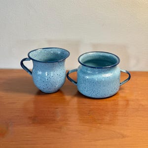 May include: Two light blue enamelware mugs with dark blue rims and handles. The mugs have a speckled pattern and sit on a wooden surface. One mug has a single handle, and the other has two.