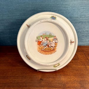 Puede incluir: Un plato de cerámica blanca vintage con un borde elevado. El centro presenta una ilustración colorida de niños en un juguete. El plato tiene el texto "The Tiny Todkins' Tearless Ride. No steed too wild for them to guide."