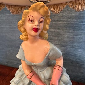 Puede incluir: Una l&aacute;mpara de mesa vintage con una figura de cer&aacute;mica de una mujer con un vestido azul y guantes rosas. La l&aacute;mpara tiene una pantalla blanca con un ribete de flecos.