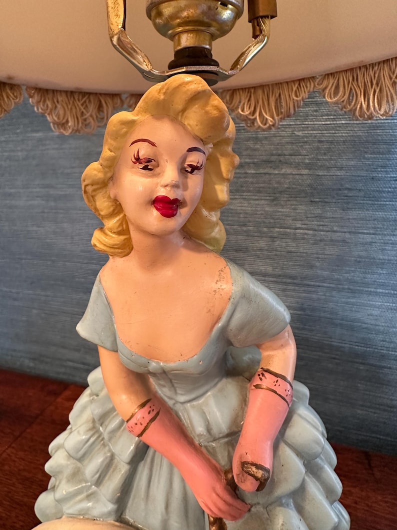 Puede incluir: Una l&aacute;mpara de mesa vintage con una figura de cer&aacute;mica de una mujer con un vestido azul y guantes rosas. La l&aacute;mpara tiene una pantalla blanca con un ribete de flecos.