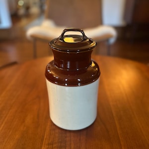 Puede incluir: Un recipiente de cerámica con cuerpo cilíndrico que presenta una base de color blanco roto con motas y una sección superior de color marrón oscuro brillante. La tapa a juego incluye un asa pequeña y arqueada en la parte superior.