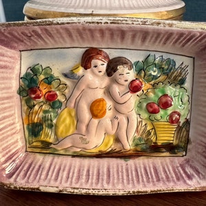 Puede incluir: Un plato de cer&aacute;mica con un borde rosa y blanco y un dise&ntilde;o en relieve de dos querubines en un jard&iacute;n. Los querubines est&aacute;n pintados en tonos de rosa, amarillo y blanco, y el jard&iacute;n est&aacute; pintado en tonos de verde, amarillo y rojo.