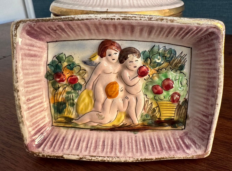 Puede incluir: Un plato de cer&aacute;mica con un borde rosa y blanco y un dise&ntilde;o en relieve de dos querubines en un jard&iacute;n. Los querubines est&aacute;n pintados en tonos de rosa, amarillo y blanco, y el jard&iacute;n est&aacute; pintado en tonos de verde, amarillo y rojo.