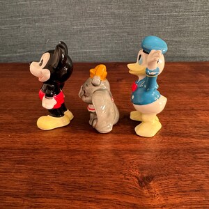 K&ouml;nnte beinhalten: Drei Keramikfiguren von Disney-Charakteren: Mickey Mouse in Schwarz, Rot und Gelb; Dumbo der Elefant in Grau und Gelb; und Donald Duck in Blau, Wei&szlig; und Gelb. Die Figuren stehen auf einer Holzoberfl&auml;che.