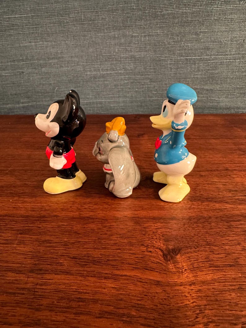 K&ouml;nnte beinhalten: Drei Keramikfiguren von Disney-Charakteren: Mickey Mouse in Schwarz, Rot und Gelb; Dumbo der Elefant in Grau und Gelb; und Donald Duck in Blau, Wei&szlig; und Gelb. Die Figuren stehen auf einer Holzoberfl&auml;che.