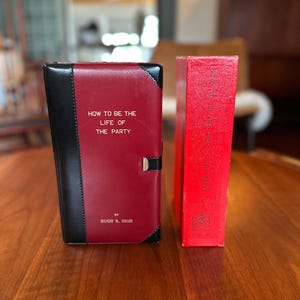 以下が含まれることがあります： ヒュー・R・ハイ著『How to Be the Life of the Party』というタイトルの本。赤と黒の表紙に金属製の留め具が付いており、背表紙にエンボス加工の文字と紋章が施された赤いスリップケースが隣に置かれています。