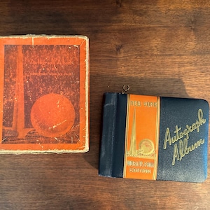 May include: Two vintage autograph albums from the New York World's Fair. One is a rectangular box with a red and orange design. The other is a black album with gold lettering and an orange stripe that reads "New York World's Fair Edition."