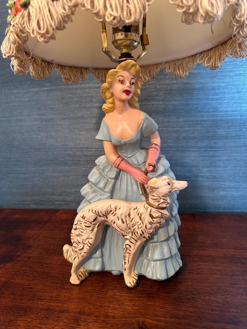 Puede incluir: Una l&aacute;mpara de mesa vintage con una pantalla blanca y una figura de una mujer con un vestido azul que sostiene un perro blanco.