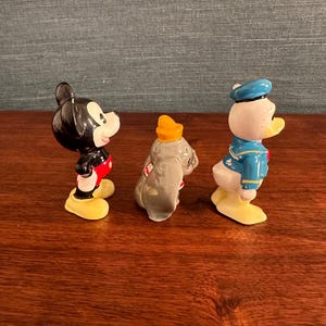 K&ouml;nnte beinhalten: Drei Keramikfiguren von Disney-Charakteren: Micky Maus, Dumbo der Elefant und Donald Duck. Micky ist schwarz mit roten Shorts und gelben Schuhen. Dumbo ist grau mit einem gelben Hut. Donald Duck ist wei&szlig; mit einem blauen Matrosenanzug und Hut.