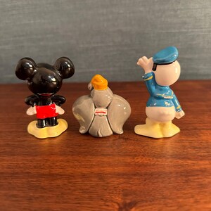 K&ouml;nnte beinhalten: Drei kleine Keramikfiguren von Disney-Charakteren. Mickey Mouse ist schwarz mit roten Shorts, einer gelben Basis. Der Dschinni ist grau mit einem gelben Turban. Donald Duck ist wei&szlig; mit einem blauen Matrosenanzug und Hut, gelbe Basis.