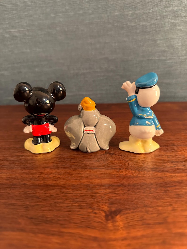 K&ouml;nnte beinhalten: Drei kleine Keramikfiguren von Disney-Charakteren. Mickey Mouse ist schwarz mit roten Shorts, einer gelben Basis. Der Dschinni ist grau mit einem gelben Turban. Donald Duck ist wei&szlig; mit einem blauen Matrosenanzug und Hut, gelbe Basis.