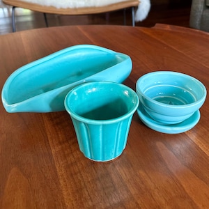 May include: A set of three turquoise ceramic items consisting of an elongated boat-shaped dish, a fluted cylindrical cup with vertical ridges, and a small round bowl with a matching saucer featuring concentric circular grooves on the interior.