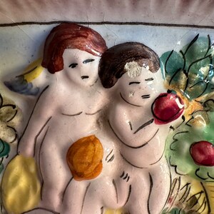 Puede incluir: Una placa de pared de cer&aacute;mica que representa a dos querubines en un entorno de jard&iacute;n. Los querubines est&aacute;n pintados en tonos de rosa y blanco, con cabello casta&ntilde;o. El jard&iacute;n presenta hojas verdes, flores amarillas y manzanas rojas.