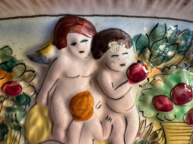Puede incluir: Una placa de pared de cer&aacute;mica que representa a dos querubines en un entorno de jard&iacute;n. Los querubines est&aacute;n pintados en tonos de rosa y blanco, con cabello casta&ntilde;o. El jard&iacute;n presenta hojas verdes, flores amarillas y manzanas rojas.
