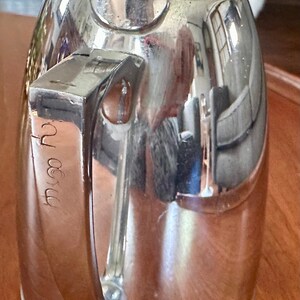 May include: A polished silver-tone metal thermal carafe featuring a hinged lid at the top and a sturdy vertical handle on the side. The handle displays engraved cursive text that appears to read 'Myra'. The surface is highly reflective.