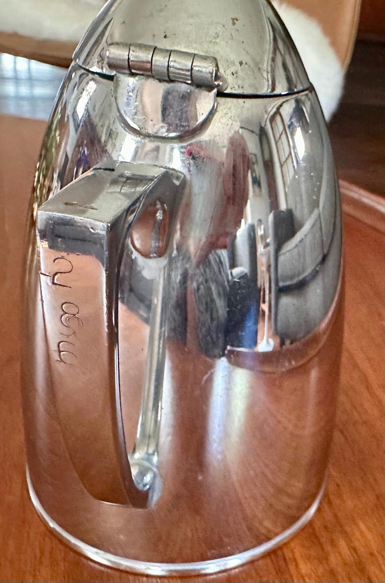 May include: A polished silver-tone metal thermal carafe featuring a hinged lid at the top and a sturdy vertical handle on the side. The handle displays engraved cursive text that appears to read 'Myra'. The surface is highly reflective.