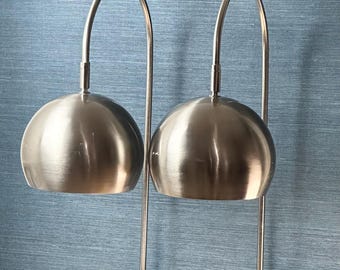 May include: Two contemporary table lamps featuring brushed metal shades and curved arms. The lamps have a spherical shade design and are set on black marble cube bases. The background is a textured blue wall.