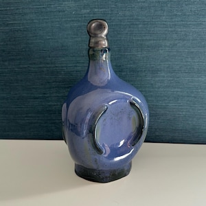 May include: A blue ceramic bottle with a silver-colored stopper. The bottle has a rounded shape with two curved indentations on the sides. The glaze has a glossy finish with variations in color.