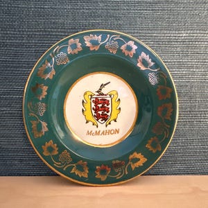 May include: A round ceramic plate featuring a dark green border decorated with gold-colored grape leaves and vines. The center is white and displays a heraldic crest with three red lions and the name MCMAHON printed in gold lettering below it.