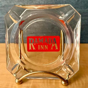 May include: Clear glass ashtray with a gold-coloured metal stand. The ashtray has a square shape with bevelled edges and a circular indentation in the centre. The red label in the centre reads "RAMADA INN" in gold lettering.