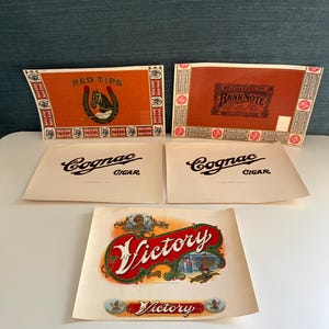 May include: A collection of vintage cigar box labels and cigar bands. The labels feature the text "RED TIPS", "BANK NOTE", "Cognac CIGAR", and "Victory". The labels are in various colors, including red, brown, and white.