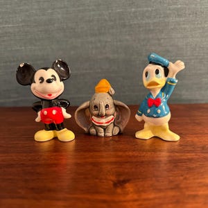 K&ouml;nnte beinhalten: Drei Disney-Keramikfiguren: Micky Maus in Schwarz, Rot und Gelb; Dumbo der Elefant in Grau, Wei&szlig; und Gelb; und Donald Duck in Blau, Gelb und Rot. Die Figuren werden auf einer Holzoberfl&auml;che pr&auml;sentiert.