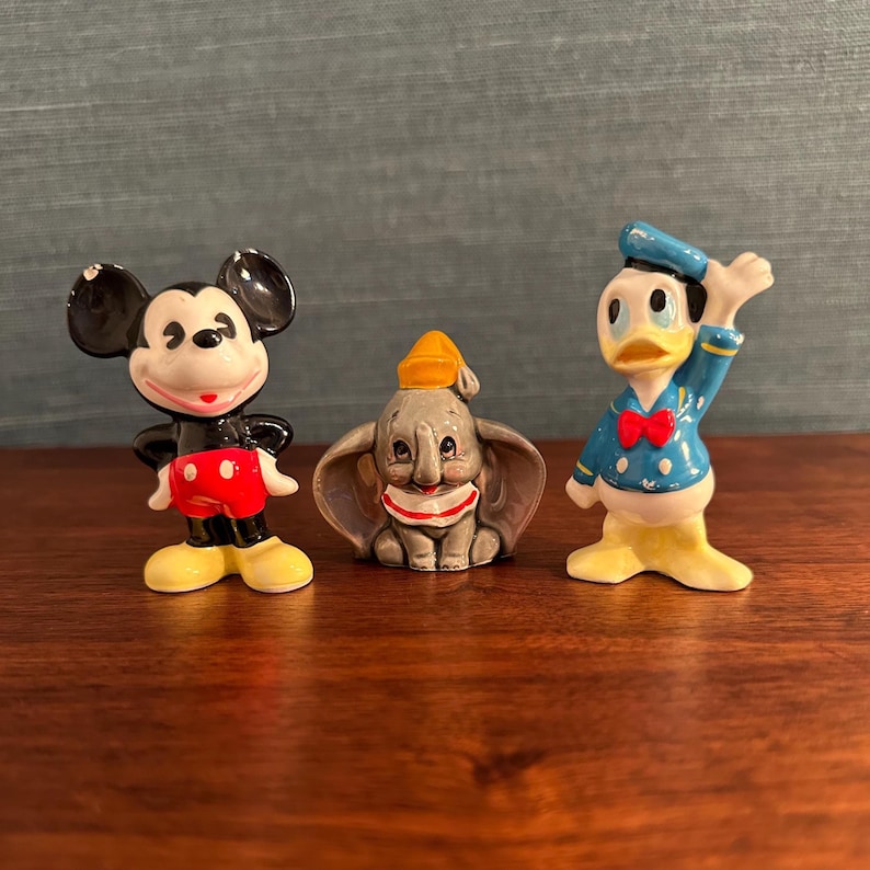 K&ouml;nnte beinhalten: Drei Disney-Keramikfiguren: Micky Maus in Schwarz, Rot und Gelb; Dumbo der Elefant in Grau, Wei&szlig; und Gelb; und Donald Duck in Blau, Gelb und Rot. Die Figuren werden auf einer Holzoberfl&auml;che pr&auml;sentiert.
