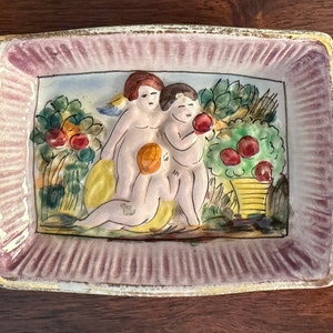 Puede incluir: Un plato de cer&aacute;mica con un borde rosa y una escena de tres querubines en un jard&iacute;n con &aacute;rboles frutales. Los querubines est&aacute;n pintados en tonos de rosa, amarillo y blanco.