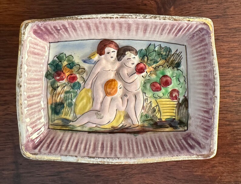 Puede incluir: Un plato de cer&aacute;mica con un borde rosa y una escena de tres querubines en un jard&iacute;n con &aacute;rboles frutales. Los querubines est&aacute;n pintados en tonos de rosa, amarillo y blanco.