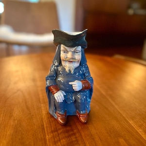 Może przedstawiać: Ceramiczny dzbanek w kształcie siedzącej postaci ubranej w ciemnoniebieski płaszcz, pasujący trójgraniasty kapelusz i czerwonobrązowe buty. Postać ma bladą cerę, cienki wąs i szpiczastą brodę, a jedna dłoń spoczywa na kolanach.