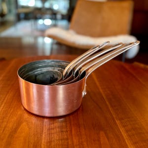 May include: A set of four nested copper saucepans with metallic handles. The pans feature a polished copper exterior and a smooth, rounded design. Each pan is stacked inside the other, with the handles extending outward in a uniform alignment.