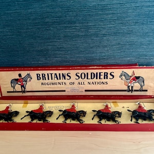 Puede incluir: Juego de juguetes vintage de Britains Soldiers, con figuras en miniatura de soldados montados con chaquetas rojas y caballos negros. Las figuras se exhiben en una caja roja con el texto "Britains Soldiers Regiments of All Nations".