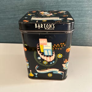 Puede incluir: Lata de caramelos Barton's negra rectangular con tapa. La lata presenta ilustraciones coloridas de cerezas, flores y cintas. El texto en la lata incluye "Barton's" y "Famous for Continental Chocolates."