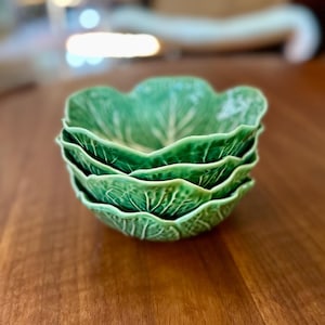 Puede incluir: Una pila de tres cuencos de cerámica verde con una superficie texturizada que imita la apariencia de hojas de col. Cada cuenco presenta un borde festoneado y ondulado, con detalles de nervaduras en relieve que irradian desde el centro.