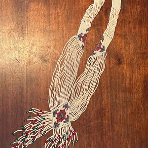 May include: Cream-coloured beaded necklace with a geometric design in red, green, and blue. The necklace has two long strands of beads with a central floral-like design and beaded fringe.