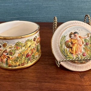 Puede incluir: Una caja de cer&aacute;mica vintage con una tapa que presenta una escena pintada a mano de dos querubines en un entorno de jard&iacute;n. La caja est&aacute; decorada con motivos florales y detalles dorados.