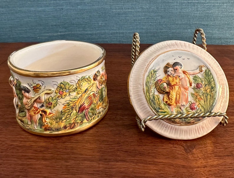 Puede incluir: Una caja de cer&aacute;mica vintage con una tapa que presenta una escena pintada a mano de dos querubines en un entorno de jard&iacute;n. La caja est&aacute; decorada con motivos florales y detalles dorados.