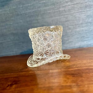 May include: A clear, glass miniature top hat with a detailed, starburst pattern. The hat has a wide brim and a cylindrical crown. The glass is transparent, allowing light to pass through, and sits on a wooden surface.