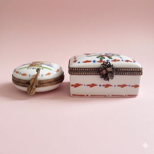 May include: Two decorative porcelain trinket boxes. One is oval with a violin charm and the word "Travail". The other is rectangular with a floral design and a bronze clasp. Both have bronze trim and are on a pink background.