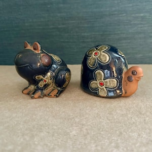 Może przedstawiać: Dwie ceramiczne figurki: żaba i żółw. Żaba jest granatowa z beżowymi akcentami i kwiatowymi detalami. Żółw jest granatowy z kwiatowymi wzorami oraz beżową głową i szyją. Ozdobne.