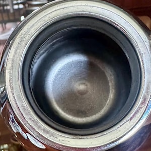 May include: A top-down view of a metallic vessel with a polished silver-toned rim and a deep, dark, matte-textured interior. The circular opening reveals a smooth, reflective metallic edge that transitions into a dark, cylindrical basin.