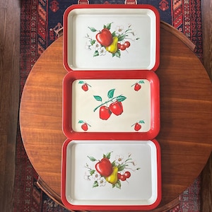 May include: Three rectangular serving trays with bright red rims and off-white centers. The top and bottom trays feature a central illustration of a red apple, a yellow pear, and small red cherries with white blossoms. The middle tray shows red apples.