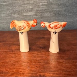 Puede incluir: Un par de saleros y pimenteros de cerámica vintage. Cada salero y pimentero tiene la forma de un pollo con cuerpo color crema, cresta roja y detalles en naranja. Los saleros y pimenteros están montados sobre un pedestal crema.