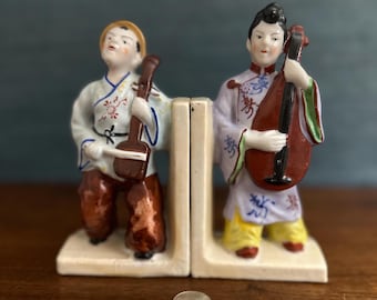 May include: A pair of vintage porcelain bookends featuring two figures in traditional Chinese clothing. The figures are playing musical instruments, one with a violin and the other with a lute. The bookends are painted in vibrant colors with intricate details.
